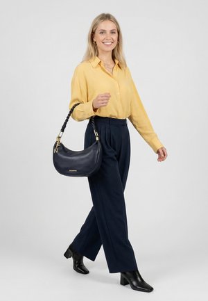 Femme marchant, portant un chemisier jaune, un pantalon plissé bleu marine, des bottes à talons noires, et portant un sac à main en cuir noir avec une bandoulière tressée.
