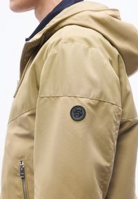 Άτομο που φοράει μπεζ μπουφάν με κουκούλα, με μαύρο κυκλικό λογότυπο στο πάνω μέρος του χεριού και μια τσέπη με φερμουάρ κοντά στη μέση.