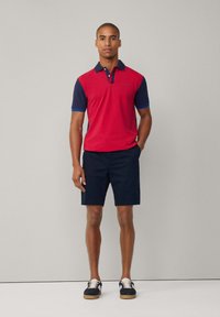 Camisa polo roja y azul marino de manga corta, combinada con pantalones cortos azul marino. Los zapatos tienen una parte superior oscura y cordones claros. Diseño simple y clásico.