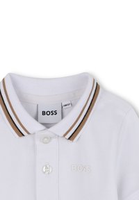 Biała koszulka polo z kołnierzykiem, z brązowym i czarnym paskiem. Wytłoczone logo "BOSS" na klatce piersiowej.