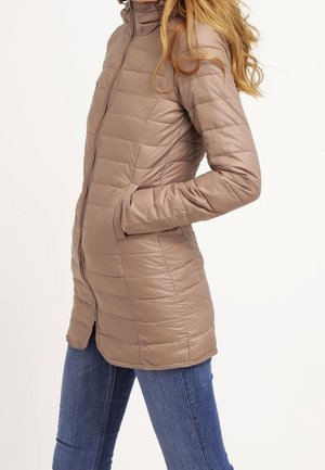 Mujer con una chaqueta larga acolchada taupe de cuello alto y bolsillos, combinada con jeans azules, de pie de lado.