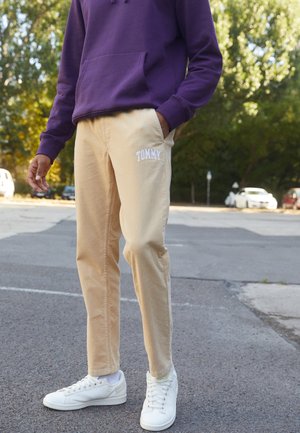 Person trägt beige Hose mit dem Schriftzug "TOMMY" und weißen Turnschuhen, steht auf einer Straße mit Bäumen und parkenden Autos im Hintergrund.