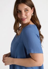 Ljusblå kortärmad topp med fyrkantig halsringning, texturerad tyg och avslappnad passform, med sömdetaljer och en frontficka.