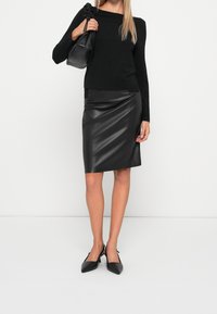 Femme portant un haut noir à manches longues, une jupe en cuir noir au genou, des chaussures à talon bas noires, tenant un sac à main noir sur l'épaule.