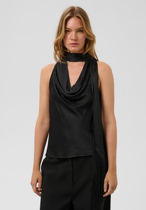 Femme portant un haut noir sans manches avec un col drapé et une longue écharpe noire sur un fond gris uni.