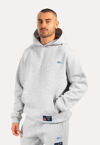 Felpa in cotone grigia con tasca frontale, logo blu sul petto e polsini a costine. Abbinata a pantaloni grigi coordinati con etichette.