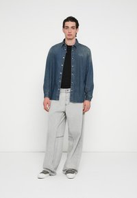 Denimskjorte i mørkeblå med knappeåpning og brodert logo, kombinert med vide grå denimbukser og grå sneakers.