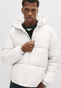 Jonge man in een witte pufferjas met capuchon, zwarte shirt eronder, zilveren ring en kettingarmband, recht vooruit kijkend met een neutrale uitdrukking.