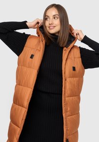 Vadderad väst i bränd orange, med huva, dragkedja och två sidofickor. Tyget har en mjuk textur med lätt glans.