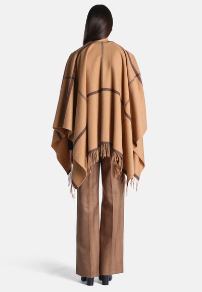 Personne vue de dos portant un poncho à franges marron clair avec un motif à carreaux foncé et un pantalon large marron assorti.