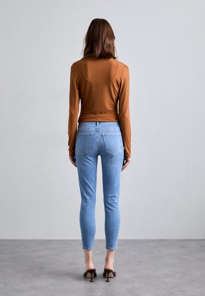 Top lungo a maniche ribbed marrone abbinato a jeans skinny blu chiaro. L'outfit è completato da mule nere con tacco e presenta una silhouette aderente.
