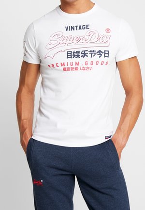 Mężczyzna w białej koszulce z logo "SuperDry" z czerwonym i niebieskim napisem oraz granatowych spodniach dresowych z czerwonym logo, ręce w kieszeniach.