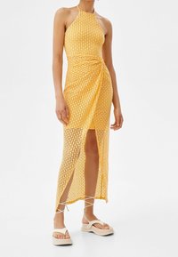 Gul virkad maxiklänning med halterneck, sidoflik och rynkdetalj vid midjan, matchad med beige platåsandaler.