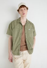 Chemise olive à manches courtes avec boutons et deux poches poitrine, portée sur un débardeur marron, complétée par une casquette beige et un pantalon crème.