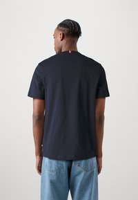 Tricou din bumbac bleu marin, cu mâneci scurte, guler rotund, având un accent mic de steag roșu, alb și albastru pe mâneca stângă, cu un croi lejer.
