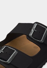 Μαύρο σανδάλι με δύο ρυθμιζόμενες μεταλλικές αγκράφες και καφέ πάτο, κοντινή λήψη σε ανοιχτό φόντο.