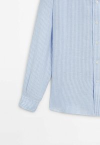 Camicia button-up di un azzurro chiaro con strisce verticali bianche, maniche lunghe, colletto classico e polsini con bottone. Il tessuto sembra liscio e leggero.