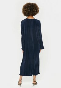 Rochie lungă plisată de culoare bleu marin, cu mâneci lungi, decolteu rotund și tiv ondulat. Textură moale, se drapează fluid până la lungimea gleznelor.