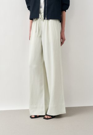 Jasnobeżowe spodnie o szerokich nogawkach ze sznurkiem w pasie, noszone z czarną krótką kurtką i czarnymi sandałami na odkryte palce na jednolitym tle.