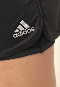 Svarta idrottsshorts med vit Adidas-logotyp som bärs av en person, som framhäver tygtexturen och den böjda nederdelen av designen.