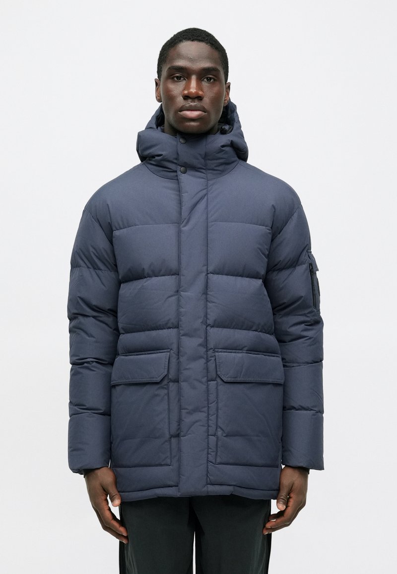 Ναυτική φούτερ τζάκετ με κουκούλα, που διαθέτει καπιτονέ σχέδιο, δύο μπροστινές τσέπες και τσέπη με φερμουάρ στον ώμο. Λείος υφαντός και ματ φινίρισμα.