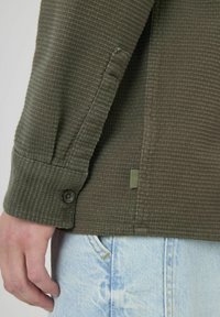 Chemise texturée vert olive avec des poignets boutonnés et un détail d'ouverture sur le côté. Tissu tissé, coupe droite, associée à un denim bleu clair.