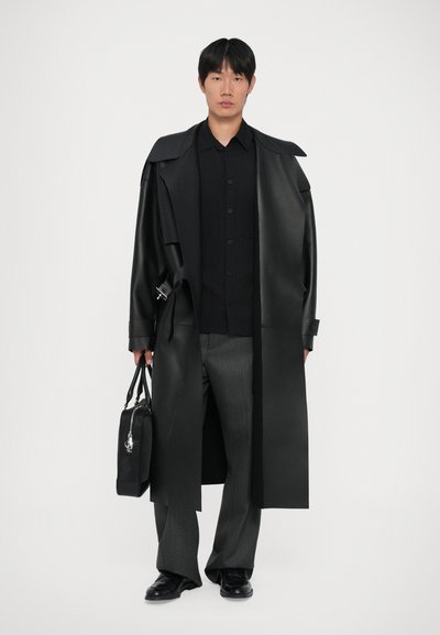 Sort læder lang frakke med oversized krave, parret med en sort button-up skjorte og grå bukser. Holder en sort taske med sølv hardware.