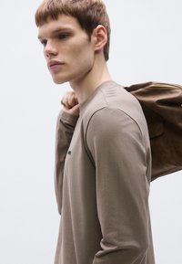 Hosszú ujjú, világosbarna ing kerek nyakkivágással. Egy barna cipzáras táska pihen a vállon. Minimalista dizájn, sima textúra.