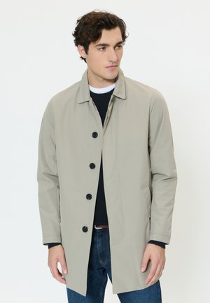 Jeune homme portant un manteau beige boutonné par-dessus un pull noir et un jean bleu, debout devant un fond blanc uni.