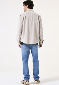 Beige overhemd met lange mouwen, relaxte pasvorm en afgeronde zoom; gecombineerd met lichtblauwe jeans en sneakers in meerdere kleuren.