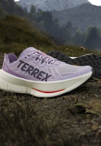Ανοιχτό μωβ παπούτσια Terrex για τρέξιμο σε μονοπάτια με λευκές σόλες, τοποθετημένα σε βραχώδες έδαφος σε ορεινό υπαίθριο περιβάλλον.