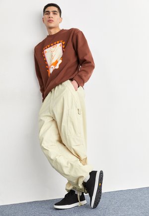 Joven apoyado en una pared blanca, viste una sudadera marrón con estampado, pantalones cargo beige y zapatillas negras con suelas blancas.