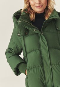 Jachetă puffer verde cu fermoar pe față, glugă mare și capse. Prezintă mansete cu nervuri și textură matlasată.