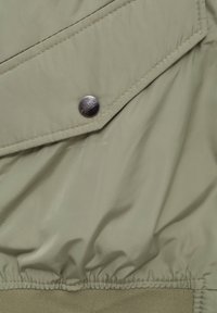 Tessuto verde chiaro, leggermente lucido, con una finitura testurizzata. Presenta un dettaglio a lembi diagonali e un bottone a pressione in metallo. Punti elastici sul fondo.