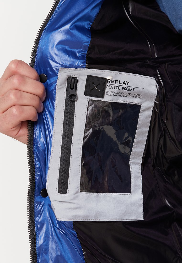 Blaue Pufferjacke mit grauem Gerätefach im Inneren, das über ein klares Fenster, einen Reißverschluss und ein Etikett mit der Aufschrift "REPLAY DEVICE POCKET" verfügt.