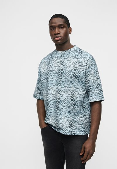 Νέος άνδρας με φαρδιά πουκάμισα με κοντά μανίκια σε σχέδιο λεοπάρδαλης και σκούρο παντελόνι, στέκεται με το ένα χέρι στην τσέπη μπροστά σε απλό φόντο.