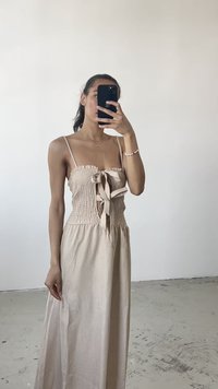 Beige, smocked dress with a fitted bodice and bow detail. Features thin straps and a flowing skirt. Smooth texture, minimalistic design.