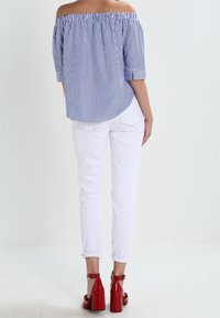 Blusa a righe blu e bianche, off-the-shoulder, abbinata a jeans skinny bianchi e sandali rossi con tacco, che mostra una vestibilità rilassata e un collo arricciato.