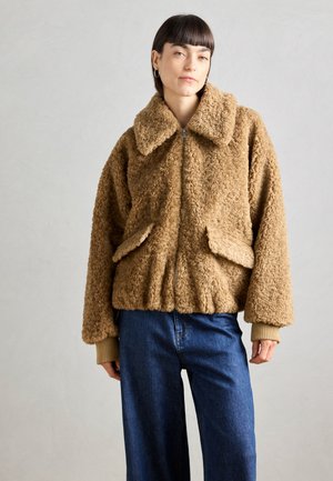 Giacca in finta pelliccia marrone con zip, grande colletto, due tasche frontali e polsini a coste. Indossata sopra jeans blu a gamba larga.