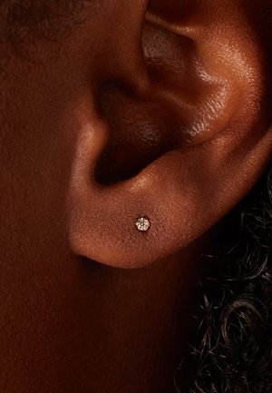 Goldfarbene Steckerohrringe mit einem kleinen, runden, klaren Kristall in der Mitte. Nahaufnahme zeigt die Textur des Ohrläppchens und die Befestigung des Ohrrings.