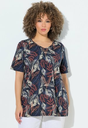 Kobieta o kręconych włosach nosi granatową bluzkę z krótkim rękawem w czerwone, białe i niebieskie wzory liści, połączoną z białymi spodniami, stojącą przy białej ścianie.