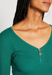 Haut vert côtelé à manches longues avec un décolleté en V profond et une petite fermeture éclair à l'avant ornée d'une tirette argentée. Texture lisse et coupe ajustée.