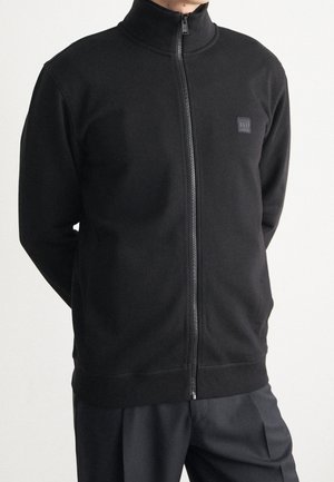 Uomo che indossa una giacca nera con zip e collo alto, con un piccolo logo quadrato sul petto a sinistra, abbinata a pantaloni neri.