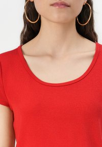 Haut rouge à manches courtes avec un col rond, fabriqué à partir d'un tissu lisse. Il possède une coupe ajustée qui met en valeur les épaules et la clavicule.