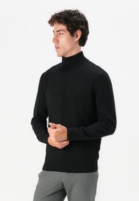 Schwarzer Rollkragenpullover mit langen Ärmeln, aus einem glatten, weichen Stoff gefertigt. Verfügt über gerippte Bündchen und Saum mit einem dezenten Logo-Detail.
