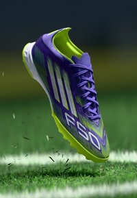 Scarpa da calcio viola e verde con un design testurizzato, strisce bianche prominenti e il marchio 'F50' in rilievo, posizionata sopra l'erba.