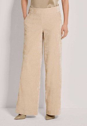 Beige weit geschnittene Hose mit Seitentaschen, getragen zu passendem beigen Oberteil und spitzen Schuhen auf neutralem Hintergrund.