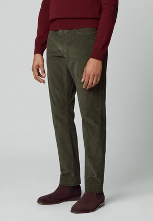 Mężczyzna noszący ciemnooliwkowe sztruksowe spodnie, bordowy sweter i pasujące bordowe zamszowe buty Chelsea, stojący na neutralnym tle.