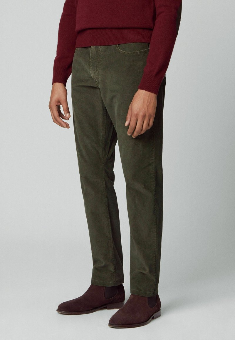 Mand iført mørke olivengrønne fløjlsbukser, bordeaux sweater og matchende bordeaux ruskind Chelsea-støvler, stående mod en neutral baggrund.