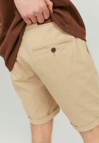 Shorts beiges avec un ourlet roulé, dotés d'une poche arrière avec un bouton marron et une texture en tissu. Une chemise marron est partiellement visible.